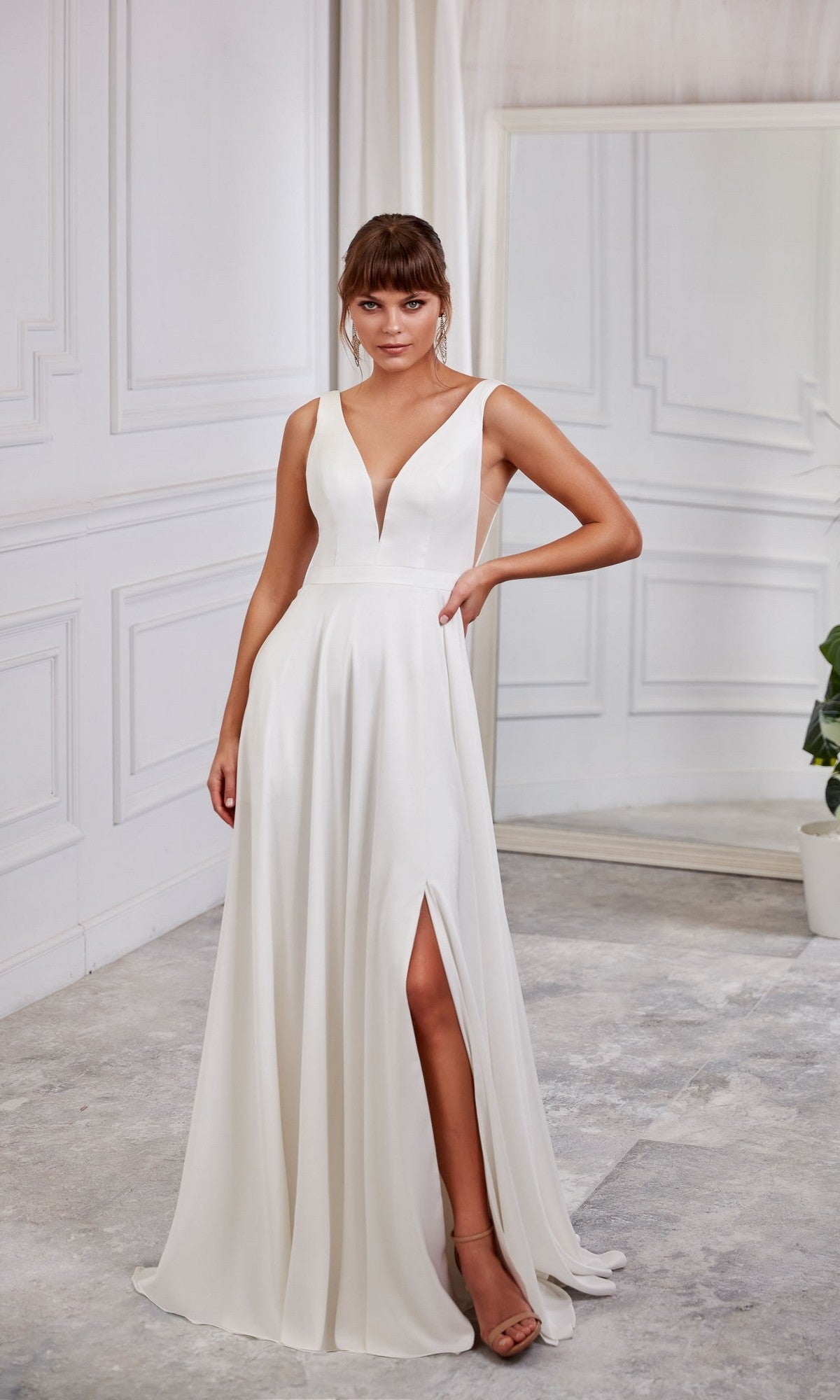 A woman poses indoors in the Aerie Couture 2007 Long White Dress, featuring a sleeveless design, deep V-neckline, and thigh-high slit. The dress is in Diamond White and she stands by a mirror and potted plant.
